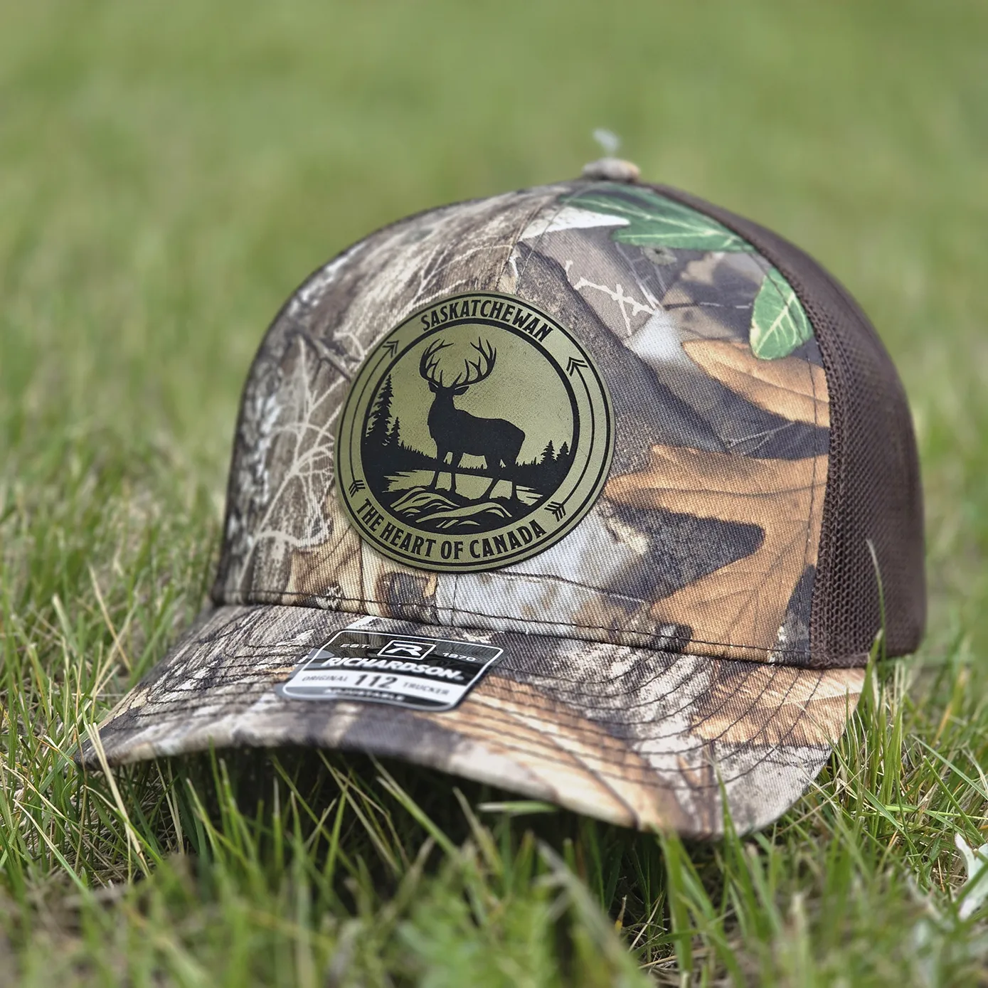 Saskatchewan The Heart of Canada Hat featuring an olive leatherette patch with a whitetail buck design on a Richardson 112 Realtree Edge camouflage trucker hat.